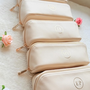 May include: A stack of cream-colored rectangular cosmetic bags with gold zippers and personalized initials in gold lettering. The bags are arranged in a row, with a few pink roses scattered around them.