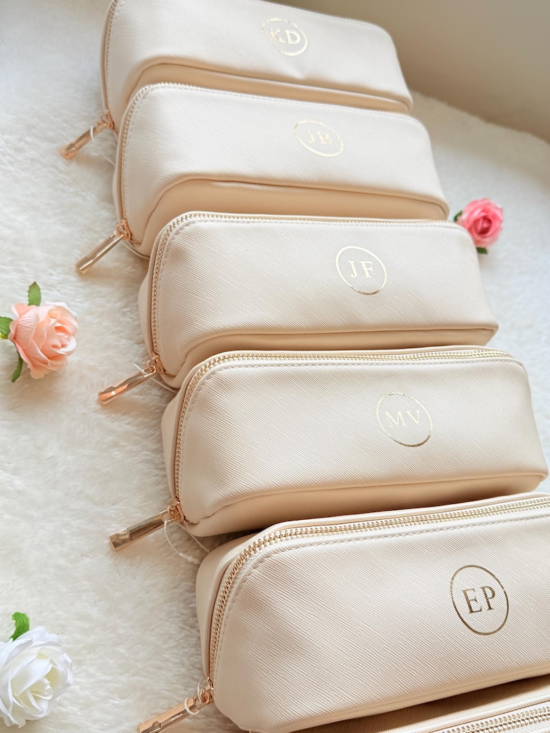 May include: A stack of cream-colored rectangular cosmetic bags with gold zippers and personalized initials in gold lettering. The bags are arranged in a row, with a few pink roses scattered around them.