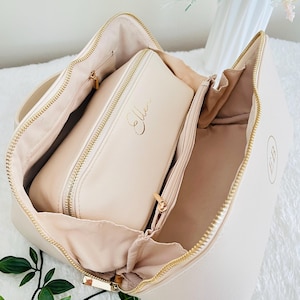 Pu&ograve; includere: Borsa per cosmetici color crema aperta con cerniere dorate e una custodia pi&ugrave; piccola abbinata all'interno. La custodia pi&ugrave; piccola ha il nome "Ellen" in oro. La borsa ha un monogramma "EB" dorato sul lato.