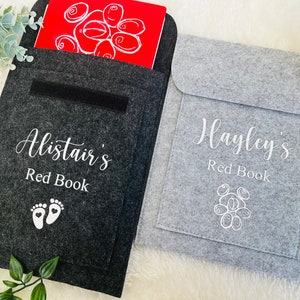 Puede incluir: Dos fundas de libro de fieltro personalizadas. La funda gris tiene el texto "Hayley's Red Book" y un contorno blanco de un racimo de uvas. La funda negra tiene el texto "Alistair's Red Book" y un contorno blanco de dos huellas con corazones.