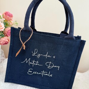 May include: Navy blue tote bag with matching handles. The bag features the text "Lynda's Match Day Essentials" in white script. A small brown tag is attached to the handle. The bag is sitting on a white surface.