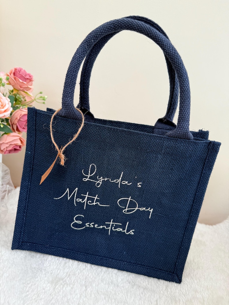 May include: Navy blue tote bag with matching handles. The bag features the text "Lynda's Match Day Essentials" in white script. A small brown tag is attached to the handle. The bag is sitting on a white surface.