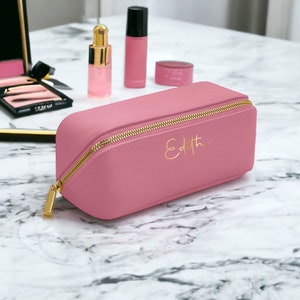Pu&ograve; includere: Un beauty case rosa con cerniera dorata e il nome "Edith" in oro. La borsa &egrave; su una superficie di marmo, con altri prodotti di bellezza sullo sfondo.