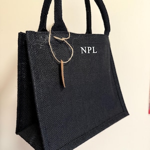 May include: A black jute tote bag with sturdy handles. The bag features the initials "NPL" in white block letters. A decorative tag hangs from the handle. The bag is designed for carrying items.