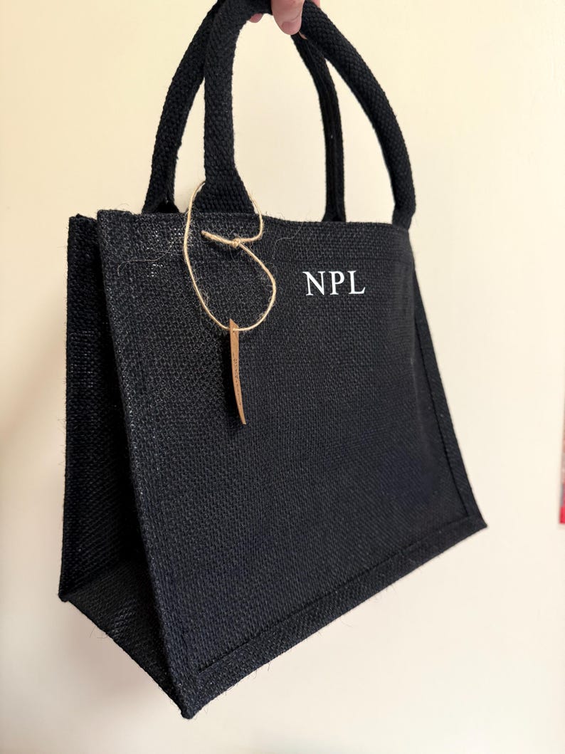 May include: A black jute tote bag with sturdy handles. The bag features the initials "NPL" in white block letters. A decorative tag hangs from the handle. The bag is designed for carrying items.
