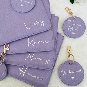 May include: A collection of lavender rectangular pouches and circular keychains with gold-tone hardware. The pouches are personalized with the names Vicky, Karen, Nanny, and Hannah in white script. The circular tags feature the text Bridesmaid and Flower Girl.