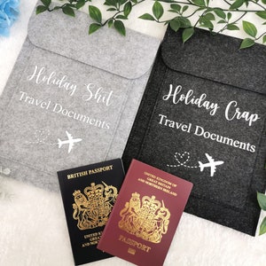 May include: Two gray felt travel document holders with the text "Holiday Shit Travel Documents" and "Holiday Crap Travel Documents" in white. Each holder features a white airplane icon with a heart-shaped trail.