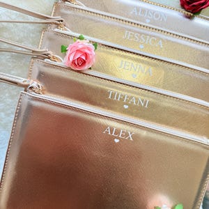 May include: A stack of five metallic rose gold wristlet pouches, each featuring a wrist strap, a zipper closure, and a personalized name printed in white with a small heart icon. The names are ALISON, JESSICA, JENNA, TIFFANI, and ALEX.
