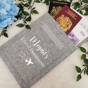 May include: Gray felt travel document holder with a white airplane and heart design. The holder has a pocket for passports and other travel documents. The text "Megan's Travel Documents" is printed on the front.