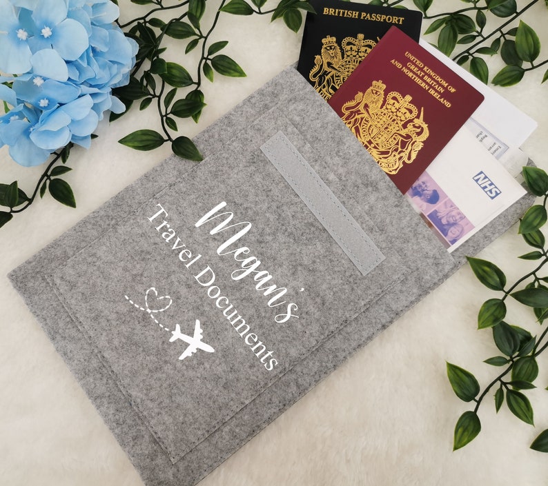 May include: Gray felt travel document holder with a white airplane and heart design. The holder has a pocket for passports and other travel documents. The text "Megan's Travel Documents" is printed on the front.