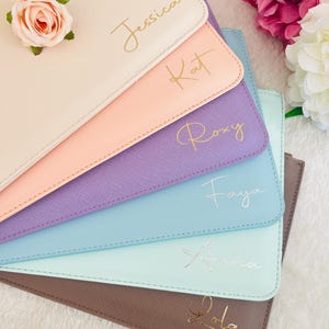 May include: A stack of six rectangular pouches in beige, peach, purple, blue, mint, and brown, each featuring a textured finish and a personalized name in cursive script. A single pink artificial rose rests on the top beige pouch.