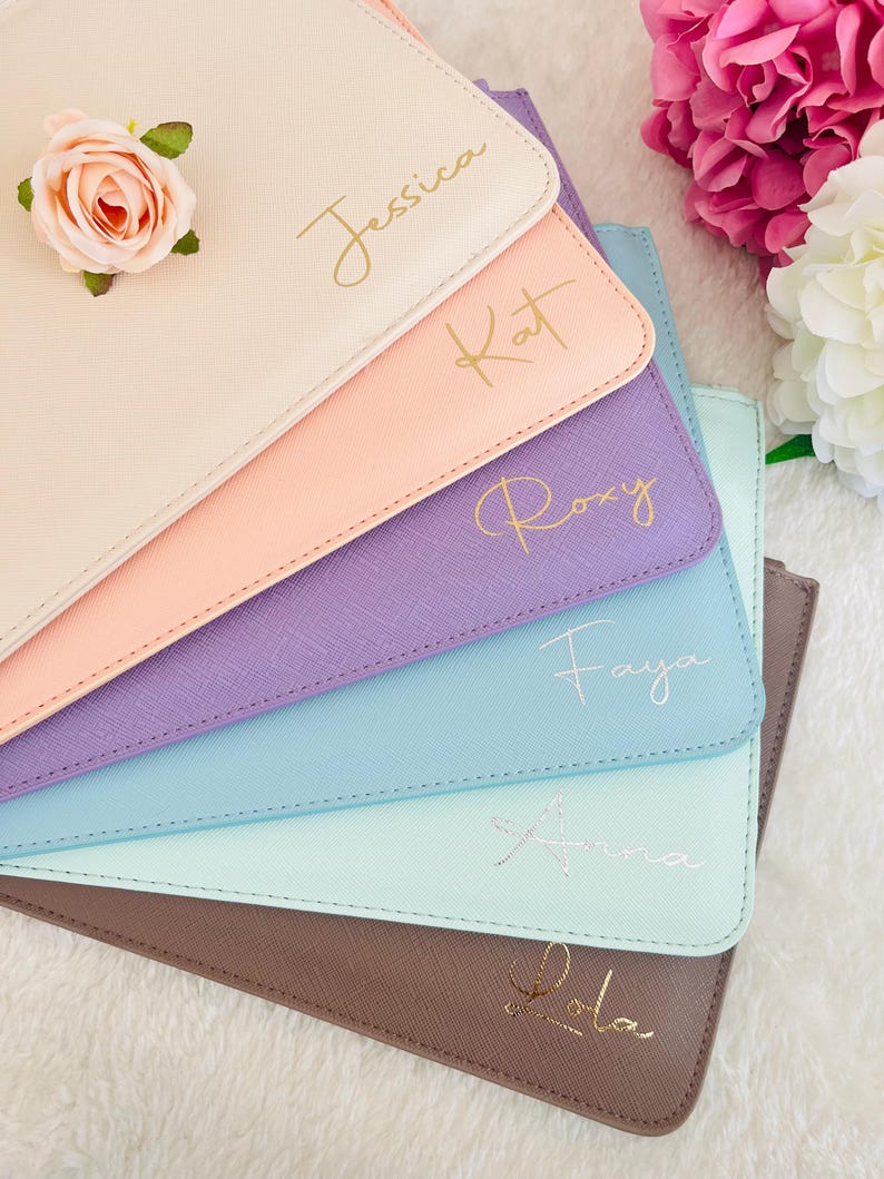 May include: A stack of six rectangular pouches in beige, peach, purple, blue, mint, and brown, each featuring a textured finish and a personalized name in cursive script. A single pink artificial rose rests on the top beige pouch.