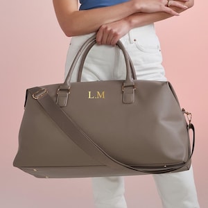 Könnte beinhalten: Eine taupefarbene Reisetasche mit zwei Tragegriffen und einem abnehmbaren Schultergurt. Die Tasche ist mit goldfarbenen Beschlägen versehen und zeigt die goldfarbenen Monogramm-Initialen L.M mittig auf der vorderen Außenfläche.