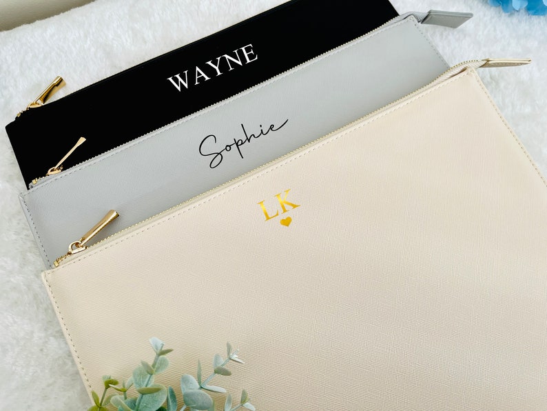 May include: Three zippered pouches in different colors and sizes. The largest pouch is cream colored with the initials "LK" in gold lettering and a small gold heart. The middle pouch is gray with the name "Sophie" written in cursive. The smallest pouch is black with the name "WAYNE" in white lettering.