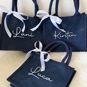 May include: Three navy blue tote bags with white cursive names: Lani, Kirsten, and Luca. Each bag has matching navy handles and a white ribbon tied in a bow. The bags are made of a textured material.