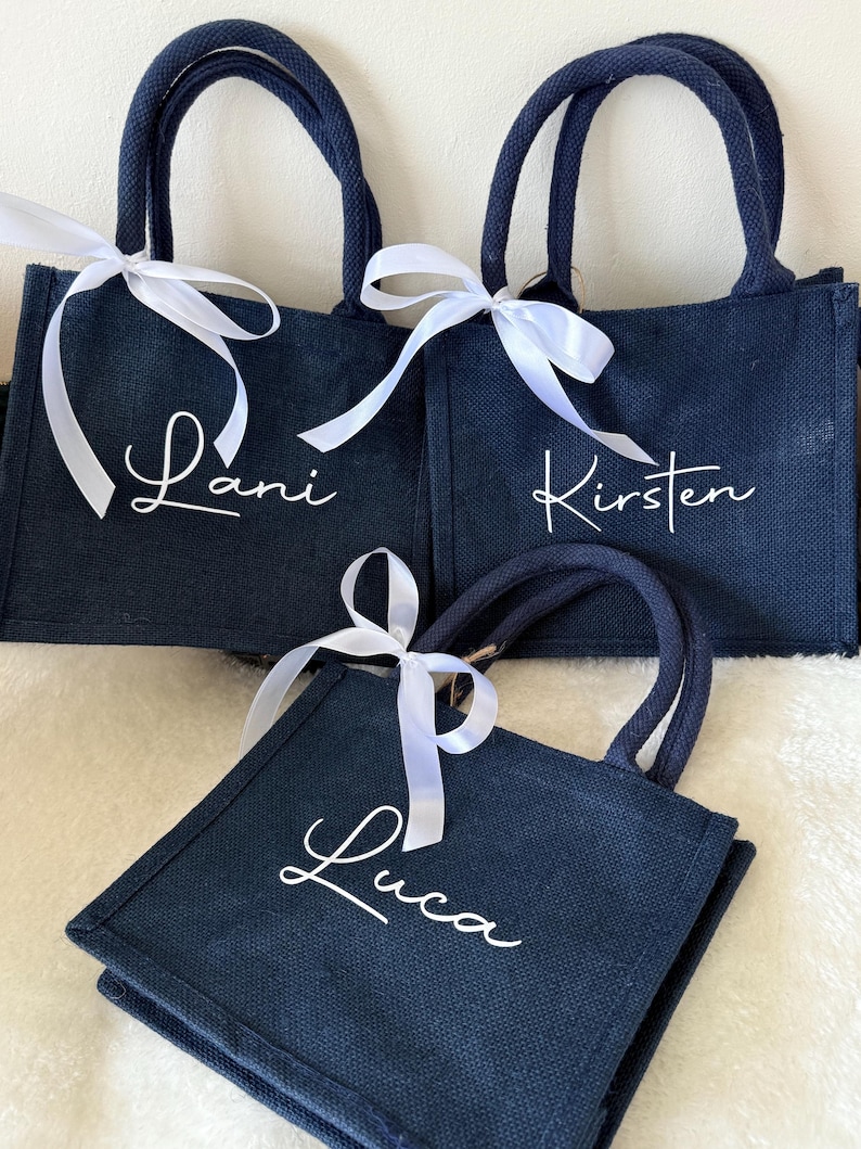 May include: Three navy blue tote bags with white cursive names: Lani, Kirsten, and Luca. Each bag has matching navy handles and a white ribbon tied in a bow. The bags are made of a textured material.