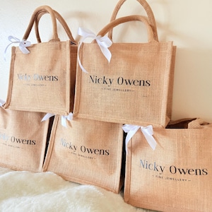May include: A stack of tan jute tote bags with brown handles and white ribbon bows. Each bag has the text "Nicky Owens - FINE JEWELLERY" printed in black. The bags are arranged on a white, fluffy surface.