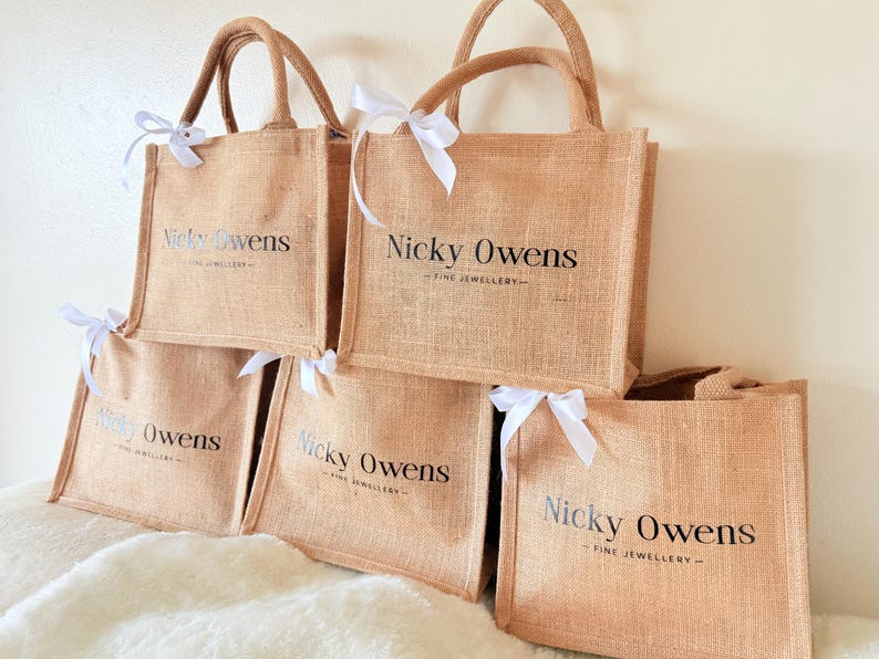 May include: A stack of tan jute tote bags with brown handles and white ribbon bows. Each bag has the text "Nicky Owens - FINE JEWELLERY" printed in black. The bags are arranged on a white, fluffy surface.