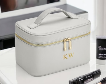 Op de afbeelding: Een grijze, rechthoekige toilettas met een gouden rits en een gouden handvat. De tas heeft de initialen "KW" in goud op de voorkant.