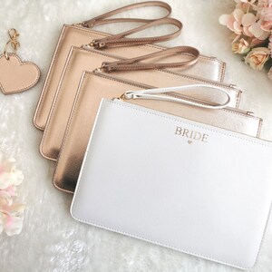 May include: Four gold clutches with a white clutch that says "Bride" in gold lettering. The clutches have gold zipper pulls and straps.