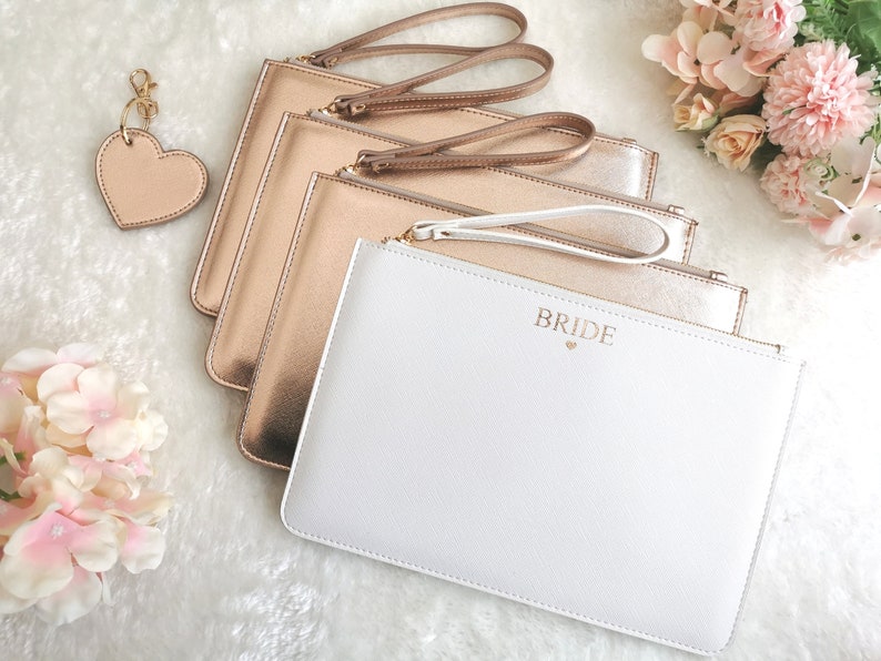 May include: Four gold clutches with a white clutch that says "Bride" in gold lettering. The clutches have gold zipper pulls and straps.