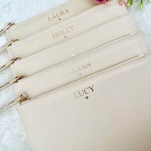 May include: Five off-white zippered pouches with gold zippers. Each pouch has a name and a small heart printed on it. The names are Laura, Holly, Lydia, Megan, and Lucy.