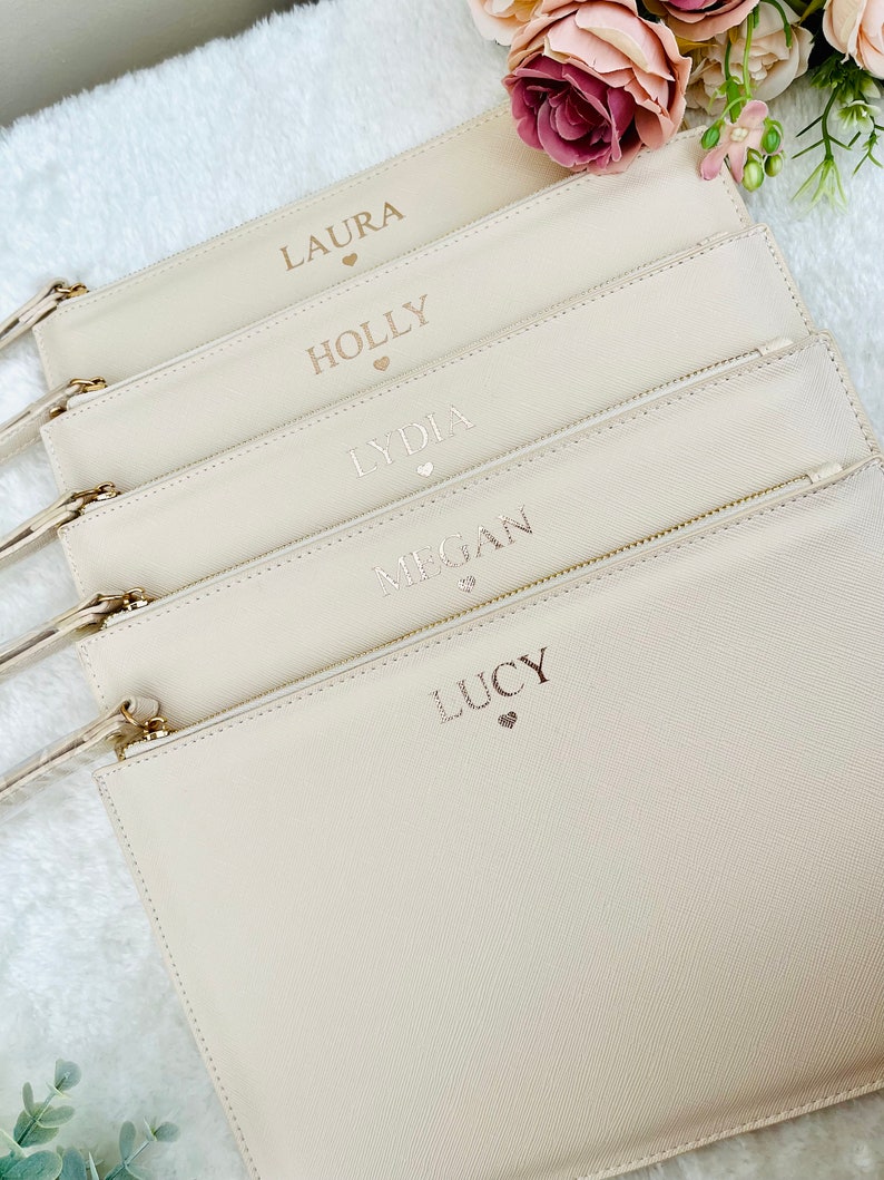 May include: Five off-white zippered pouches with gold zippers. Each pouch has a name and a small heart printed on it. The names are Laura, Holly, Lydia, Megan, and Lucy.