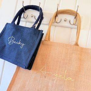 May include: Two small tote bags, one navy blue and one peach, hang from hooks. The navy bag has "Becky" embroidered in gold script, while the peach bag has "Angela" embroidered in gold script. Both bags have matching handles.