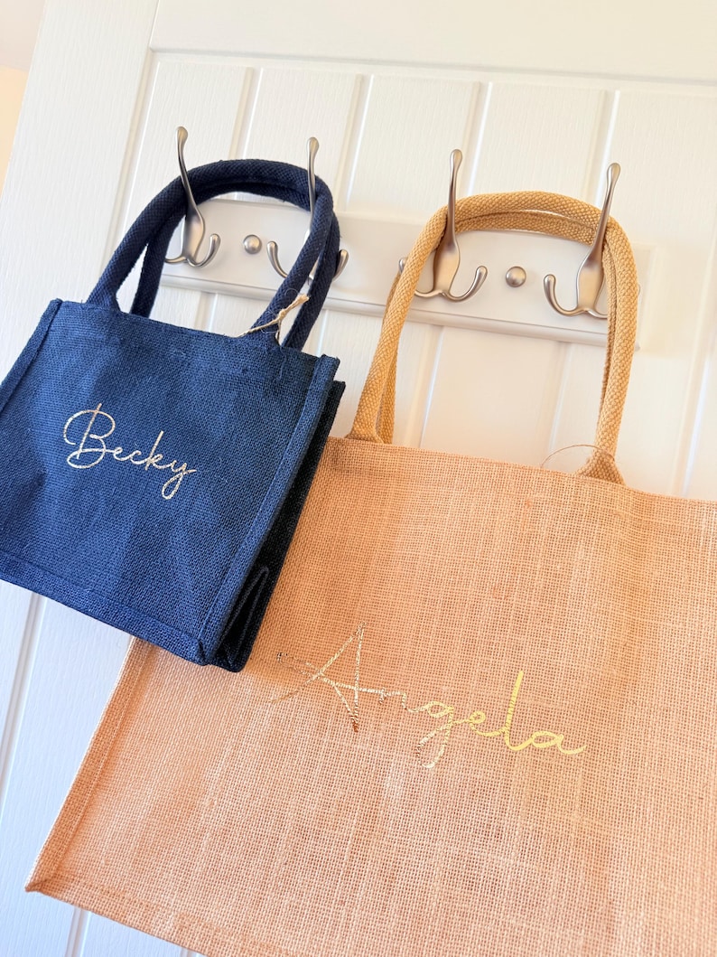 May include: Two small tote bags, one navy blue and one peach, hang from hooks. The navy bag has "Becky" embroidered in gold script, while the peach bag has "Angela" embroidered in gold script. Both bags have matching handles.