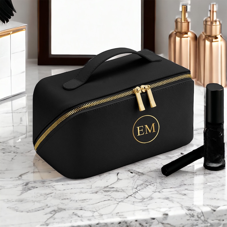May include: Black rectangular makeup bag with a gold zipper and handle. The bag has a gold initial monogram in a circle. A black nail polish bottle and applicator are next to the bag.