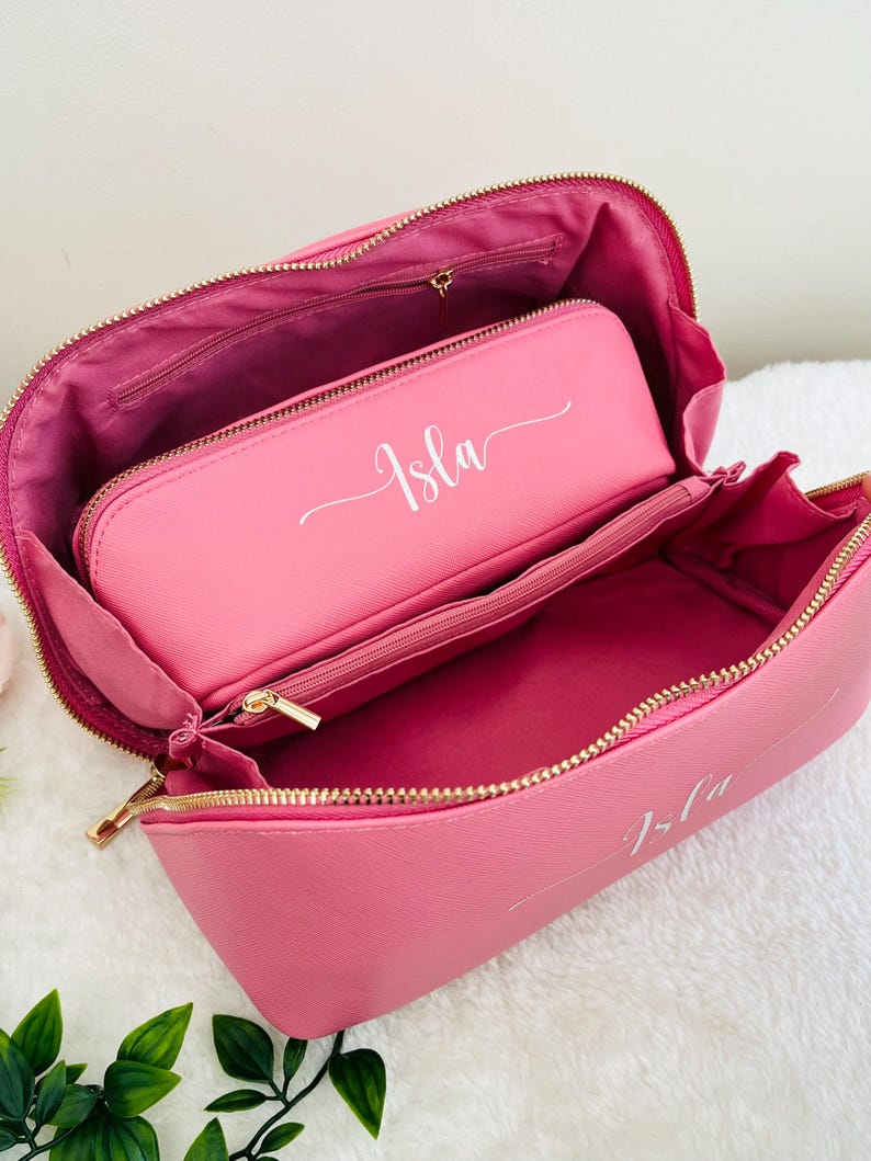 Pu&ograve; includere: Borsetta per cosmetici rosa aperta, con una pochette pi&ugrave; piccola abbinata all'interno. Entrambi i pezzi hanno cerniere dorate e il nome "Isla" in corsivo bianco. La borsa &egrave; aperta, rivelando la fodera interna e pi&ugrave; scomparti.