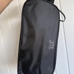 May include: A black rectangular zippered storage bag with a smooth fabric exterior and a black handle at the top. The bag features a white embroidered monogram with the letters H and E in a cursive script style on the lower right side.