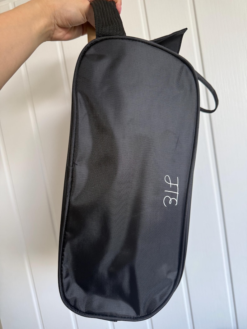 May include: A black rectangular zippered storage bag with a smooth fabric exterior and a black handle at the top. The bag features a white embroidered monogram with the letters H and E in a cursive script style on the lower right side.