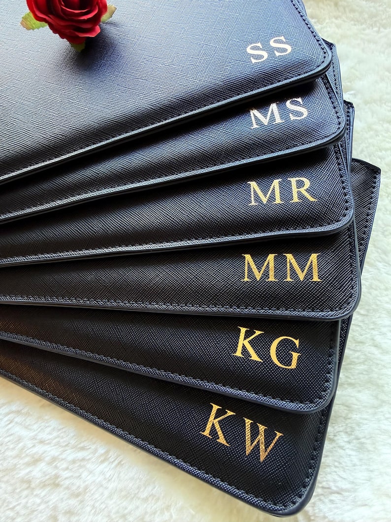 May include: A stack of black rectangular pouches with a textured finish, each featuring gold-colored embossed initials including SS, MS, MR, MM, KG, and KW. A single red rose is placed on the top corner of the uppermost pouch.