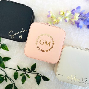 K&ouml;nnte beinhalten: Drei kleine quadratische Schmuckk&auml;stchen mit verschiedenen personalisierten Designs. Die erste Box ist schwarz mit dem Namen "Sarah" und einem Herzen. Die zweite Box ist rosa mit einem goldenen Kranz und den Initialen "GM". Die dritte Box ist wei&szlig; mit dem Namen "Abbie" und einem Herzen.