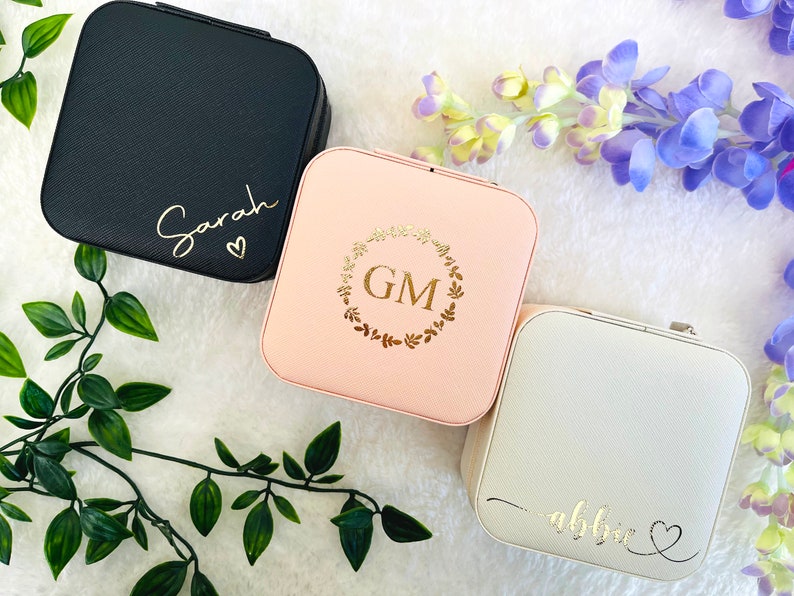 K&ouml;nnte beinhalten: Drei kleine quadratische Schmuckk&auml;stchen mit verschiedenen personalisierten Designs. Die erste Box ist schwarz mit dem Namen "Sarah" und einem Herzen. Die zweite Box ist rosa mit einem goldenen Kranz und den Initialen "GM". Die dritte Box ist wei&szlig; mit dem Namen "Abbie" und einem Herzen.