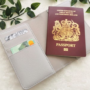 May include: A gray passport holder with two card slots. The holder is open and a British passport is visible inside. The passport is red with gold lettering and a royal crest.