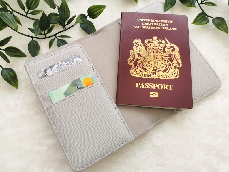 May include: A gray passport holder with two card slots. The holder is open and a British passport is visible inside. The passport is red with gold lettering and a royal crest.