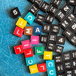 May include: A collection of square game tiles featuring black tiles with white alphanumeric codes and colorful tiles in red, yellow, green, blue, pink, and brown, each displaying a single white letter or number, scattered across a blue surface.