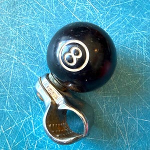May include: A black billiard ball with a white number eight inside a white circle, attached to a silver-colored metal clamp. The clamp features a circular opening and visible metallic texture with signs of wear and oxidation on the inner surface.
