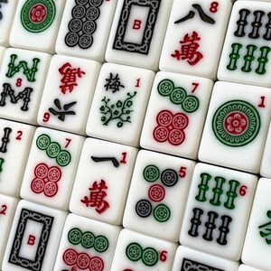 May include: A close-up of a set of mahjong tiles. The tiles are white with black and green characters and symbols. Some of the tiles have red dots. The tiles are arranged in a grid pattern.