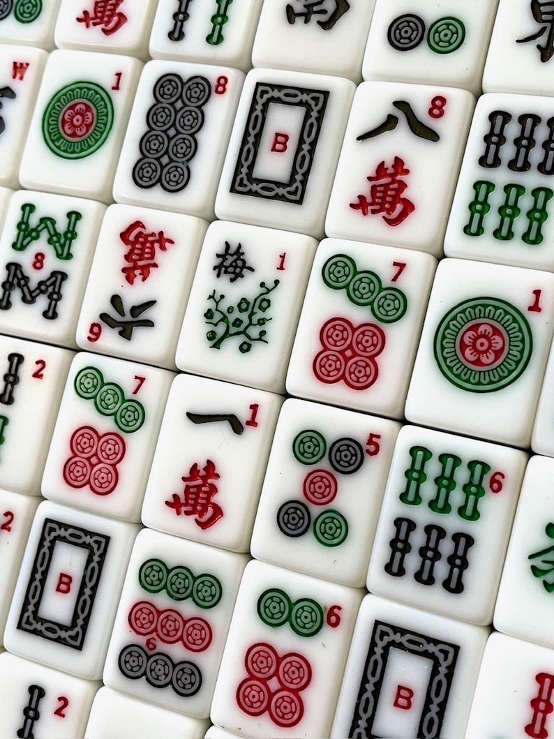 May include: A close-up of a set of mahjong tiles. The tiles are white with black and green characters and symbols. Some of the tiles have red dots. The tiles are arranged in a grid pattern.