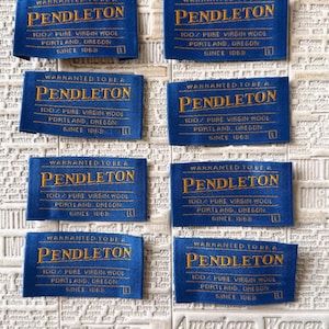 May include: Multiple blue Pendleton labels with gold text are arranged on a vintage newspaper background. Each label reads "WARRANTED TO BE A PENDLETON 100% PURE VIRGIN WOOL PORTLAND, OREGON SINCE 1863".
