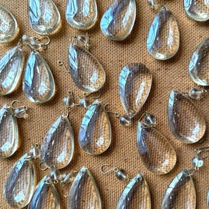 May include: A collection of clear, teardrop-shaped glass crystals, each featuring a smooth, polished surface and a faceted edge. Every crystal is attached to a small, circular glass bead and a thin metal wire hook for hanging purposes.