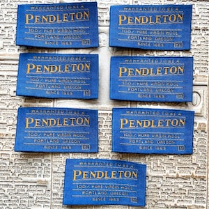 May include: Seven rectangular blue fabric labels with gold embroidered text. Each label reads Warranted to be a Pendleton, 100% Pure Virgin Wool, Portland Oregon, Since 1863, with size indicators M or XL printed in the bottom right corner.