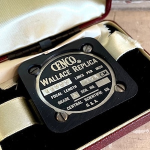 May include: A vintage black Cenco Wallace Replica scientific instrument, displayed in an open burgundy case with a cream satin lining. The instrument has silver text that reads "Focal Length" and "Lines per Inch". The case is open, revealing a black velvet interior.