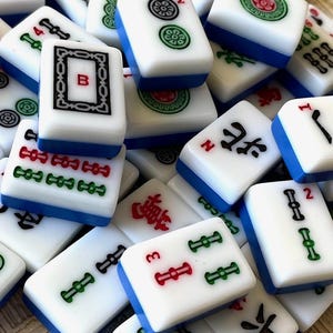 May include: A close-up of a collection of white and blue mahjong tiles. The tiles feature various Chinese characters, numbers, and symbols. Some tiles have red and green accents.