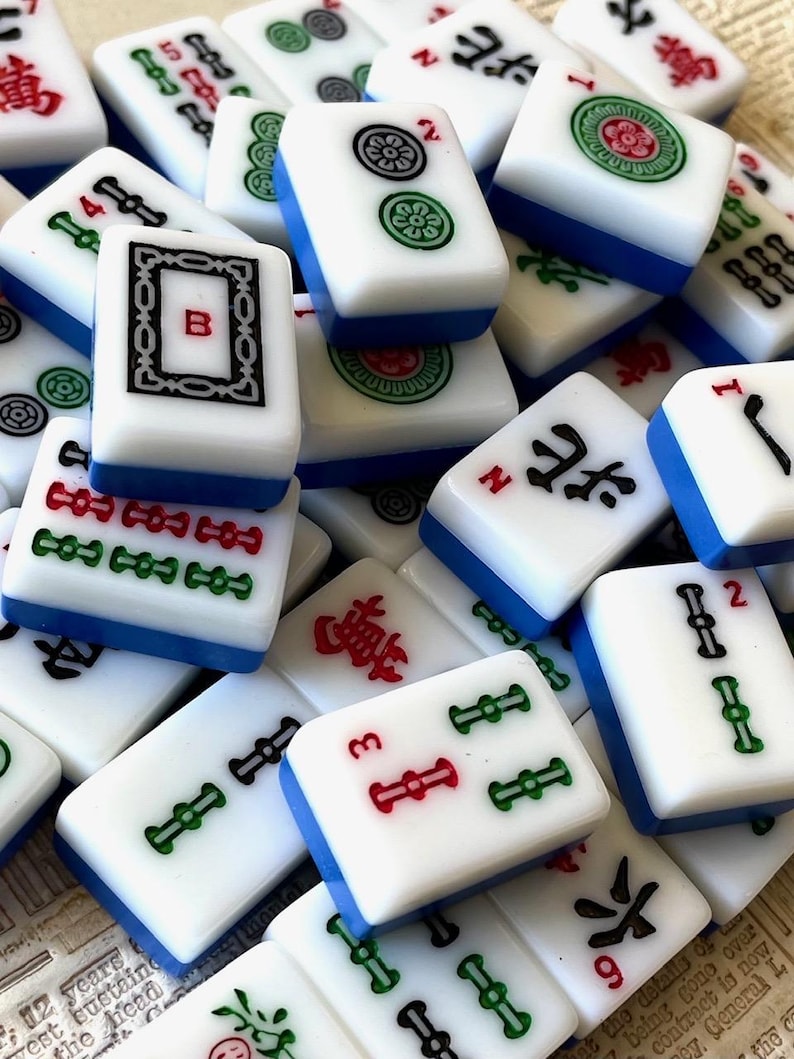 May include: A close-up of a collection of white and blue mahjong tiles. The tiles feature various Chinese characters, numbers, and symbols. Some tiles have red and green accents.
