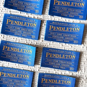 May include: Several blue Pendleton labels with gold text are arranged on a newspaper background. Each label reads "WARRANTED TO BE A PENDLETON 100% PURE VIRGIN WOOL PORTLAND, OREGON SINCE 1863".