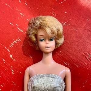 May include: A vintage doll with blonde hair and blue eyes. The doll is wearing a silver strapless top. The background is a distressed red surface.
